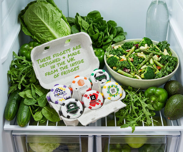 Branded fidget toys in a cardboard egg carton inside a refrigerator filled with fresh produce, illustrating CHX's UK supply chain story from end-of-life fridges to manufactured products